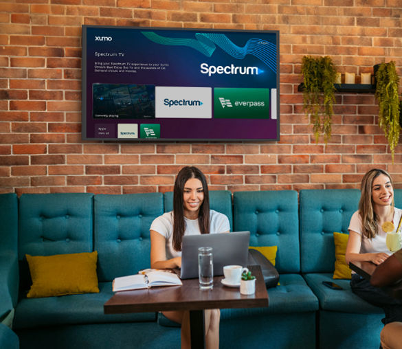 Women sitting at a table with a laptop and notebook, TV in the background showing Spectrum streaming options