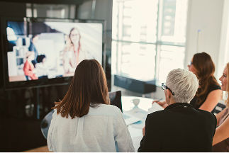 A group of women in an office, sitting around a glass table looking at a television